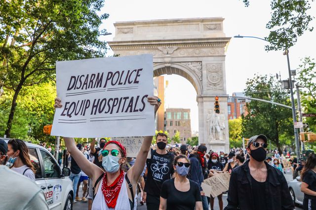 Protesters in masks march from Washington Square Park with the arch in the background; one is holding a sign that says "Disarm Police, Equip Hospitals"